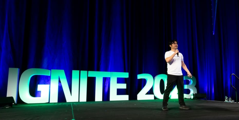 An Asian man wearing a white t-shirt is standing on a stage in front of a sign reading IGNITE 2023. 