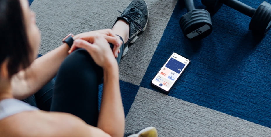 a woman wearing athletic gear is sitting on the floor looking at a health app on her phone. There are hand weights next to her.  